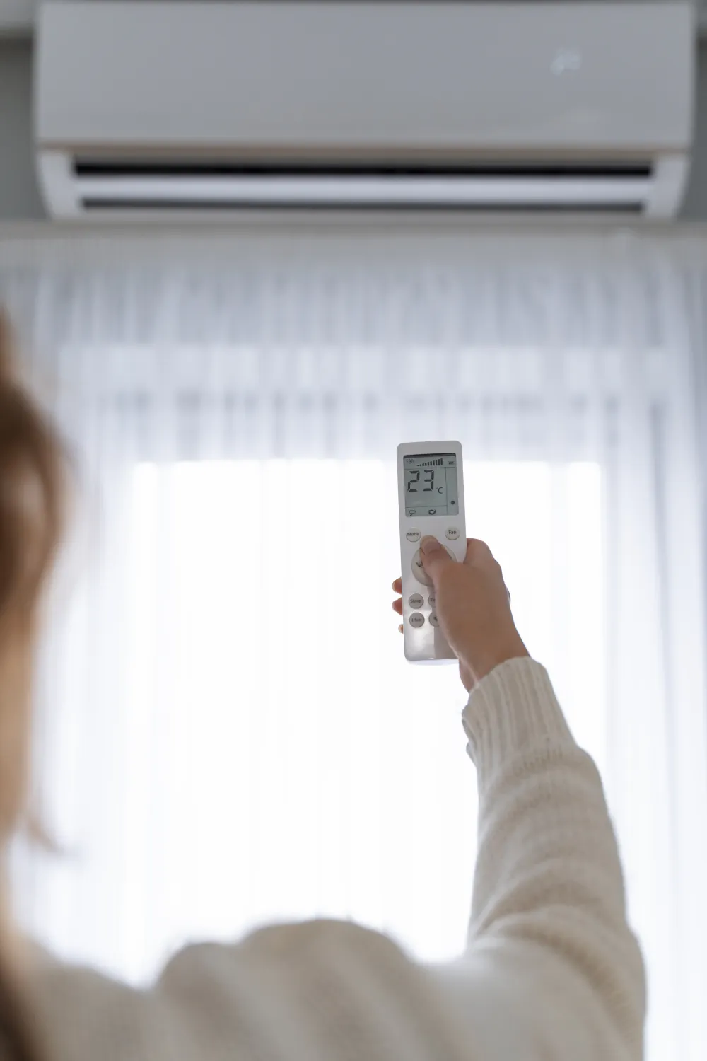 Femme réglant sa climatisation à 23 °C devant une fenêtre : malgré la clim, la chaleur du soleil entre par le vitrage. Le film solaire PrivTeint la bloque à la source, à Hyères et dans le Var.