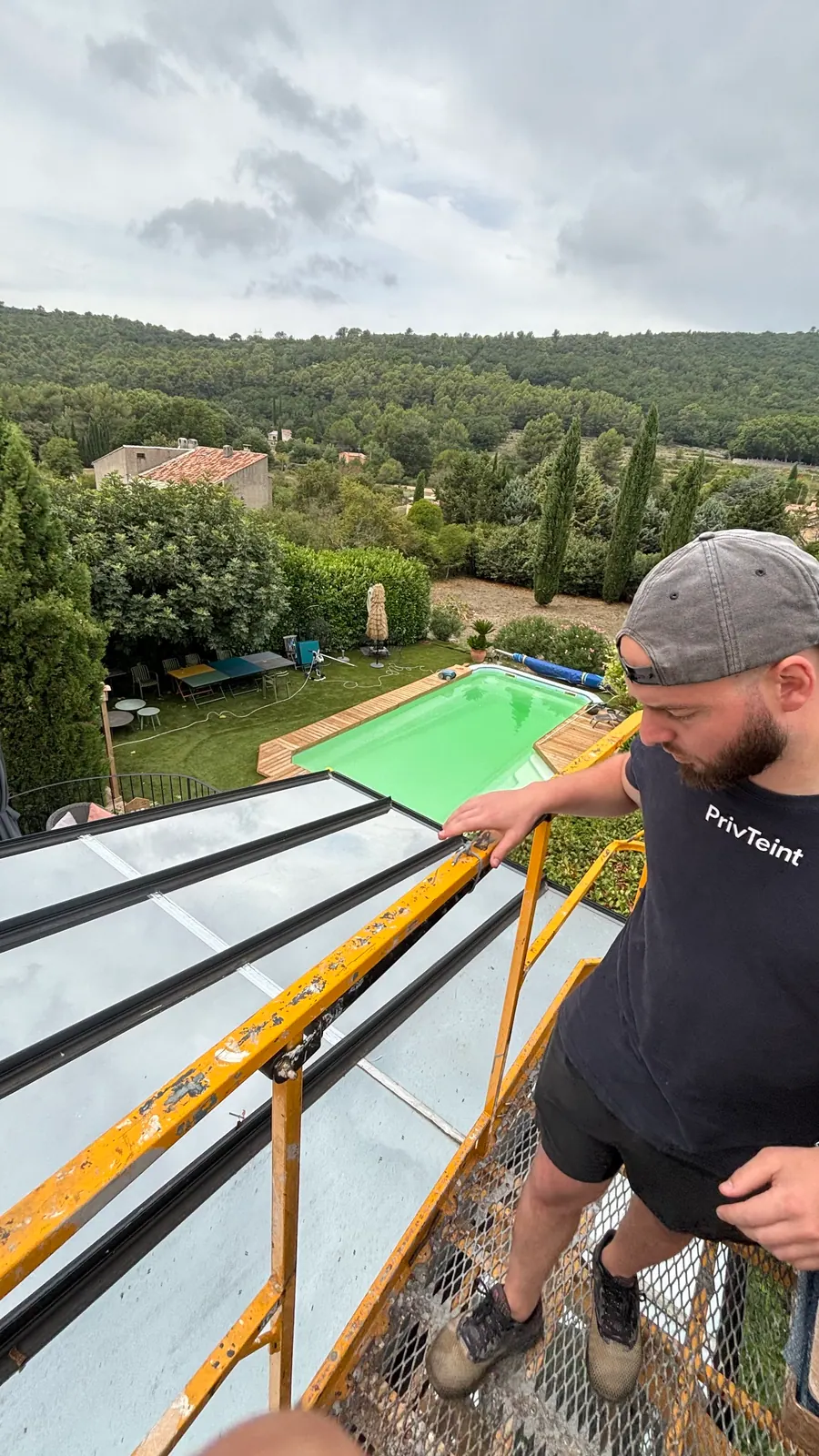 Technicien PrivTeint travaillant depuis le panier de la nacelle au-dessus de la toiture vitrée, jardin et piscine en contrebas